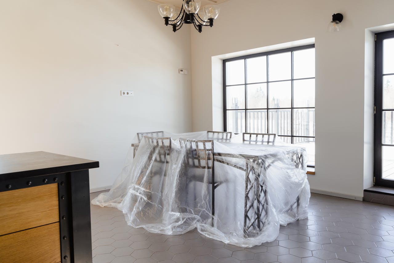 Bright dining room with table and chairs wrapped in protective plastic for renovation.